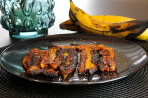 Fried plantains served on a black plate with fresh bananas in the background.